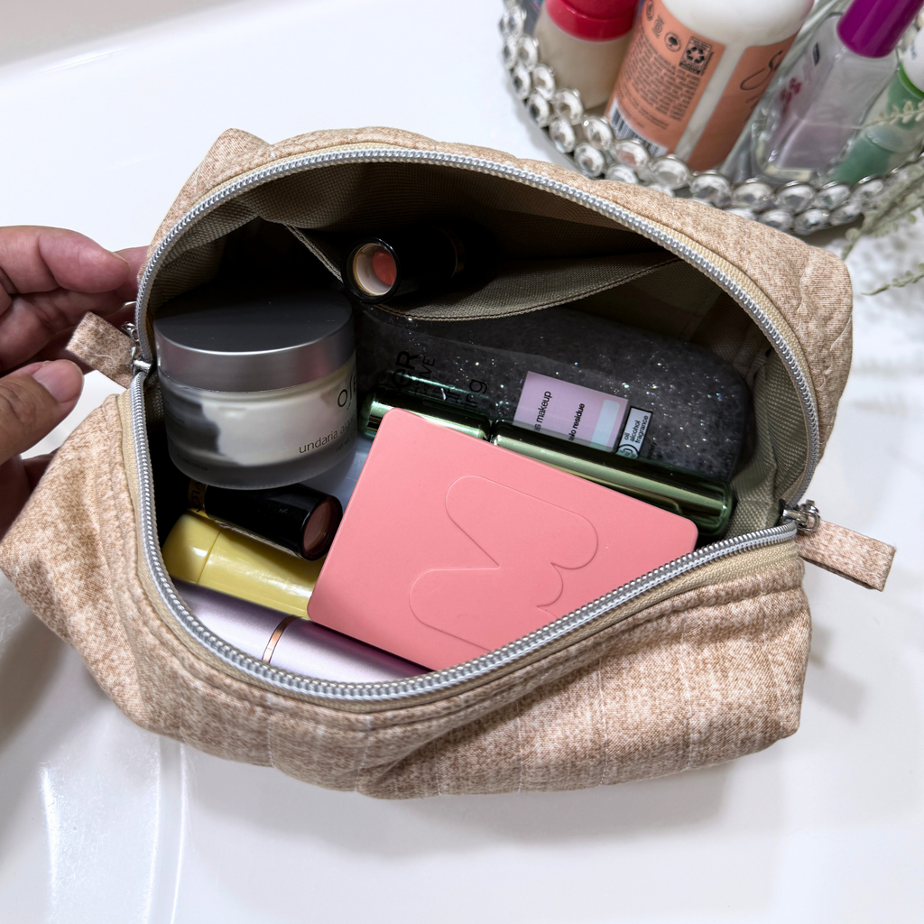 A beige quilted cosmetic pouch with a silver zipper, shown open and filled with various makeup items. A hand is holding the left side of the bag, and a bathroom sink and toiletry tray are visible in the background.
