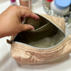 A close-up of a hand opening a beige textured cosmetic pouch with a silver zipper, showing the interior lining, with toiletry bottles blurred in the background.