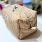A beige textured fabric cosmetic pouch with a silver zipper, shown on a white surface with a blurred background.