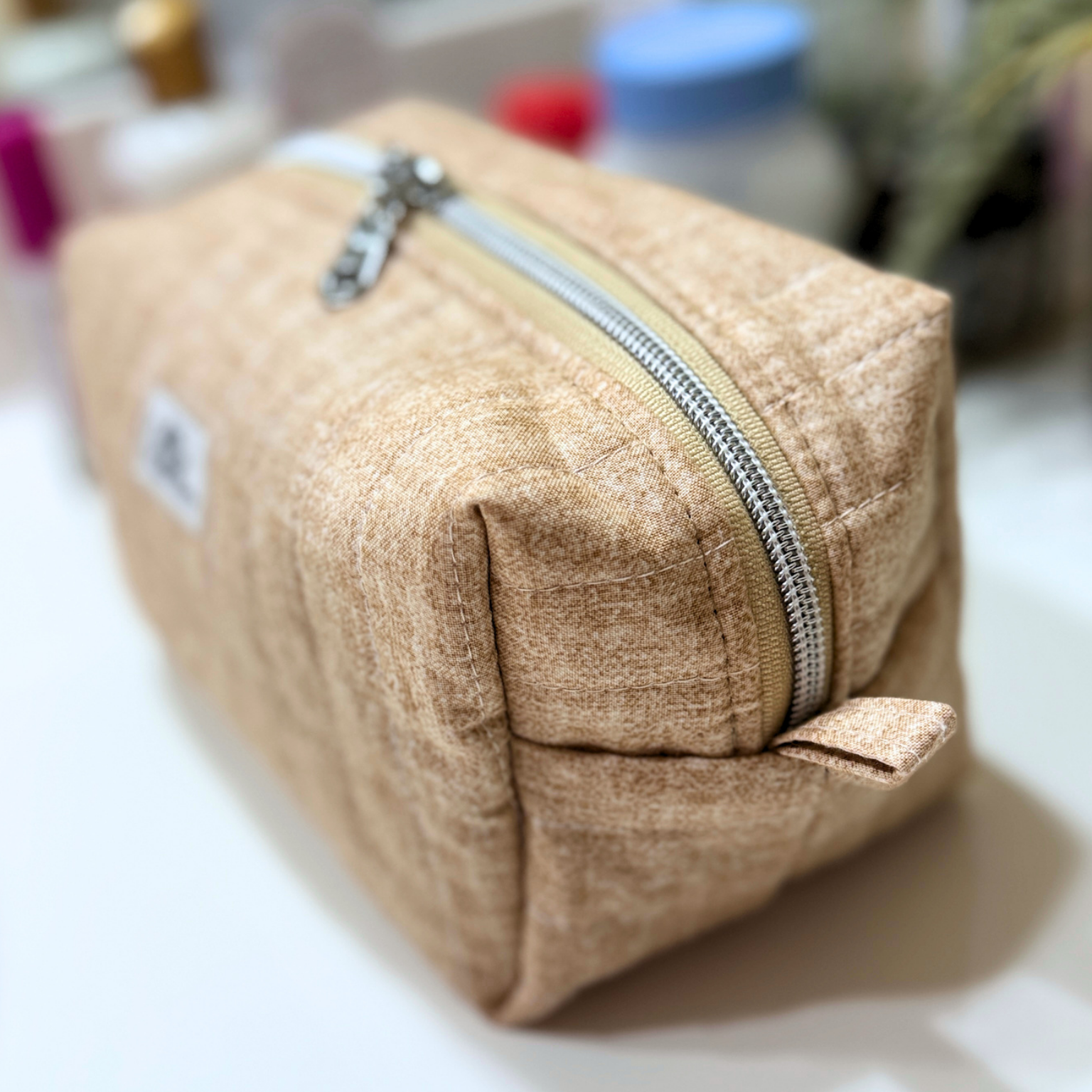 A beige textured fabric cosmetic pouch with a silver zipper, shown on a white surface with a blurred background.