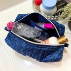 A blue denim-style zippered cosmetic pouch filled with toiletries, including a clear bottle with a pink cap and a pink-capped item, resting on a white surface with blurred bottles and greenery in the background.