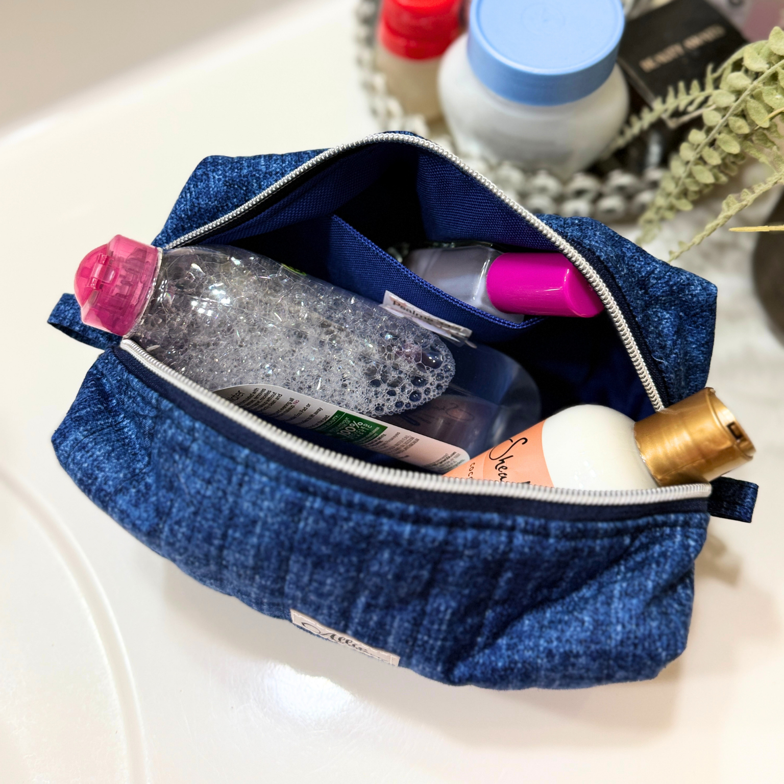 A blue denim-style zippered cosmetic pouch filled with toiletries, including a clear bottle with a pink cap and a pink-capped item, resting on a white surface with blurred bottles and greenery in the background.