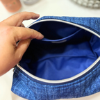 A close-up of a blue textured zippered pouch being opened by a hand, showing a bright blue interior lining with a silver zipper.