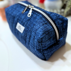 A blue quilted fabric zippered pouch with a silver zipper and a small white label, photographed on a white surface with a softly blurred background.