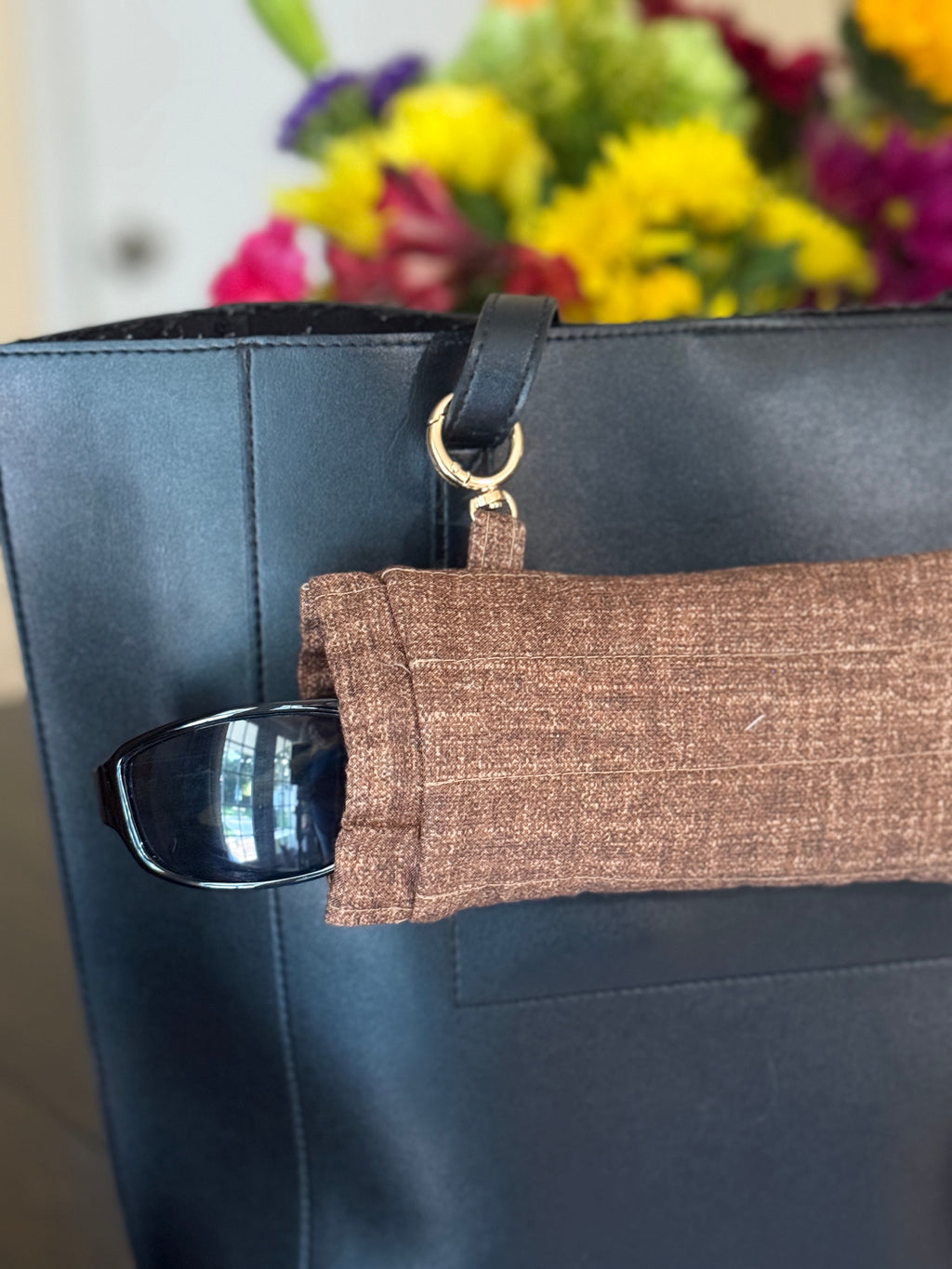 A close-up of a black tote bag with a brown woven fabric sleeve holding a pair of sunglasses, with a colorful blurred flower arrangement in the background.