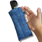 A hand holding a blue denim-textured sunglasses pouch with a black pair of sunglasses inside, on a white background.