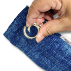 A close-up of a hand holding a silver metal key ring attached to a textured blue fabric pouch or sleeve against a white background.
