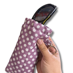 A hand holding a lavender fabric sunglasses pouch with a white floral pattern, containing black sunglasses with a clear case, against a white background.
