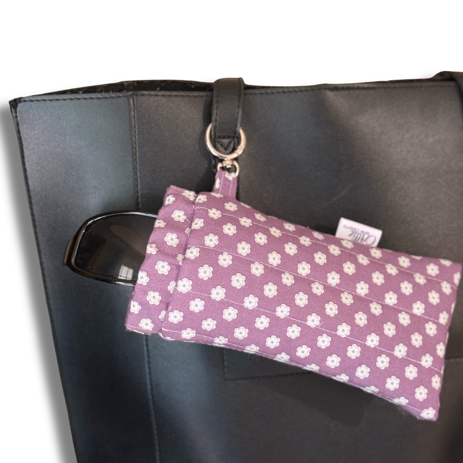 A purple fabric pouch with a white floral pattern hangs from a black tote bag using a black strap and silver metal ring. A pair of black sunglasses is tucked into the side pocket of the tote bag.