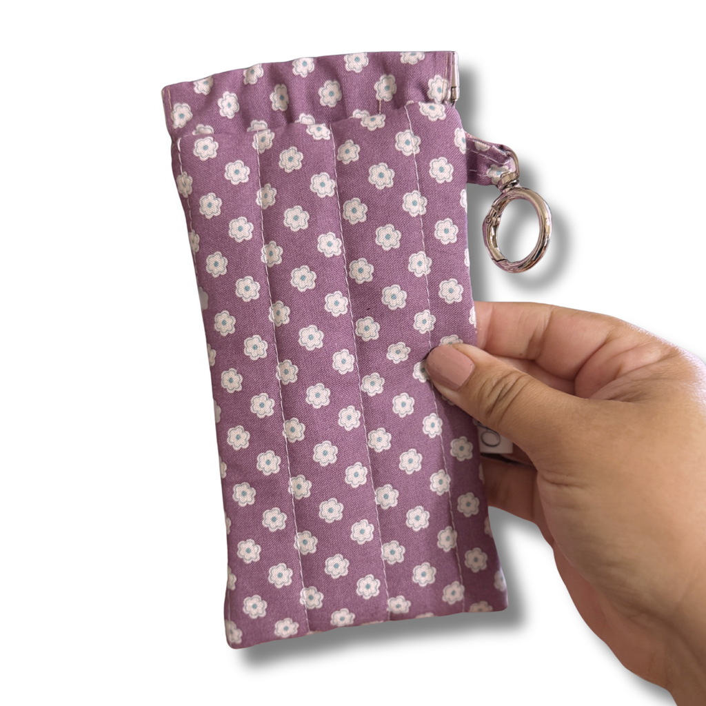 A hand holding a small fabric pouch with a purple and white floral pattern, featuring a metal key ring attachment, set against a plain white background.