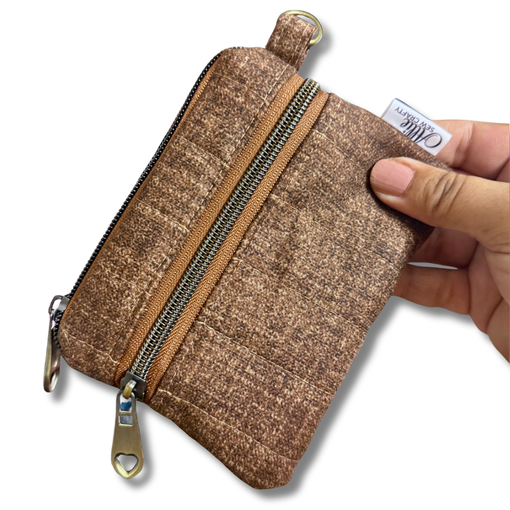 A hand holding a small brown textured zippered pouch with a gold-tone zipper and pull tab, shown against a plain white background.