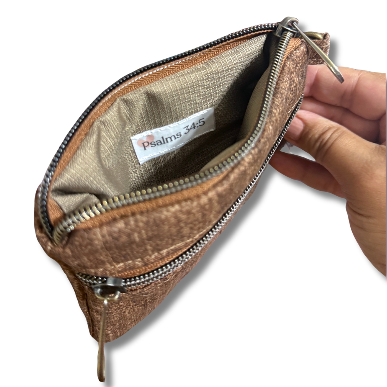 A small zippered pouch with a textured metallic bronze-brown exterior and a beige interior lining. A hand is holding the open pouch to show the inside, which features a white label reading 'Psalms 34:5'.