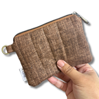 A hand holding a small brown quilted fabric pouch with a zipper closure, a metal ring, and a small fabric tag, shown against a plain white background.