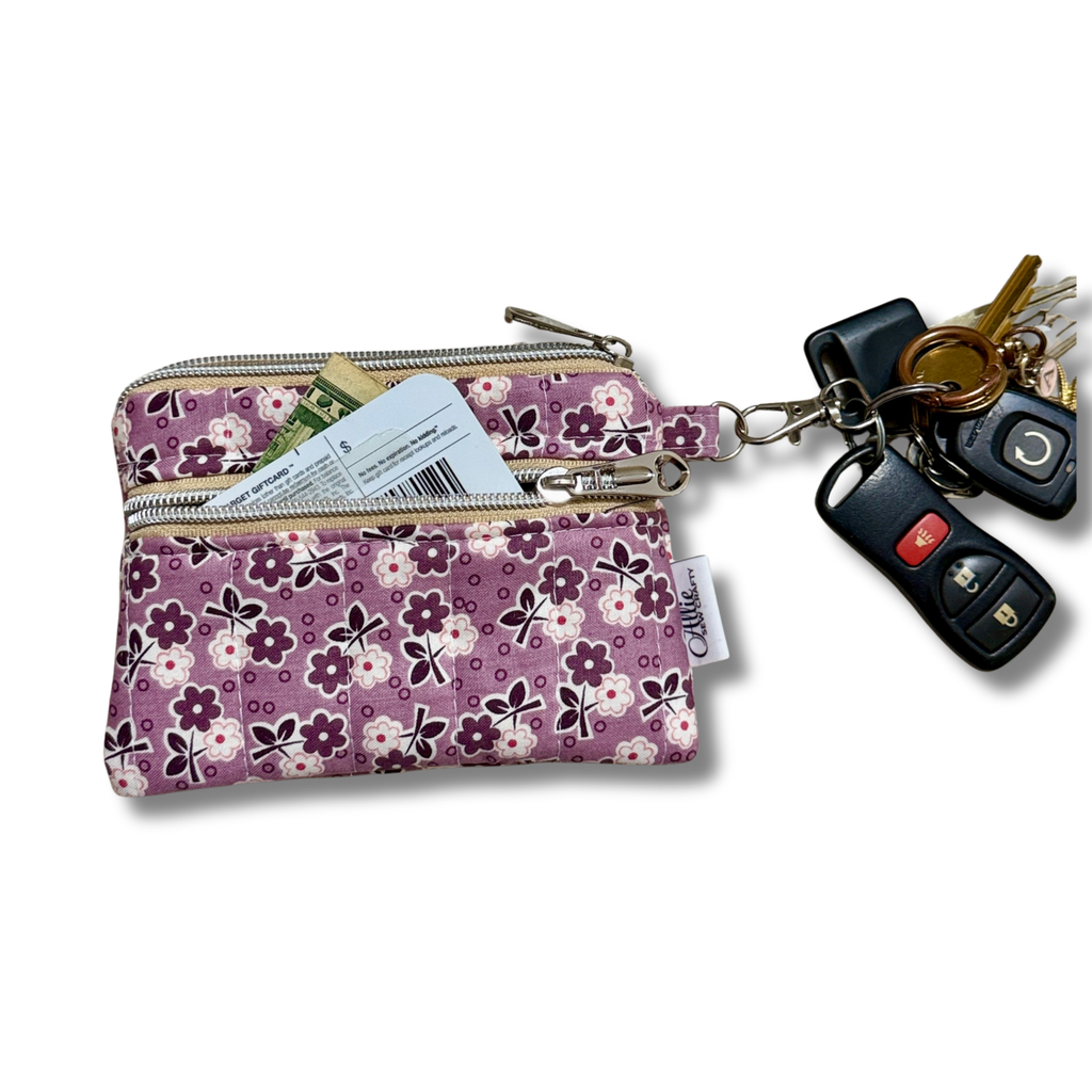 A small floral zippered pouch in purple, pink, and white, shown with keys and a credit card tucked into the top zipper. A small fabric tag on the right side reads 'Allie Sew Crafty'.
