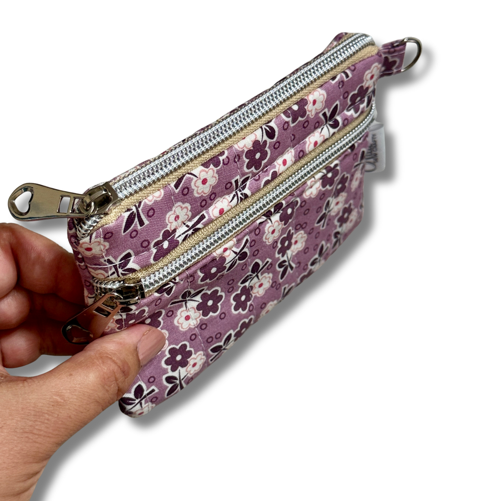 A hand holding a small zippered pouch with a purple floral print, shown against a plain white background.