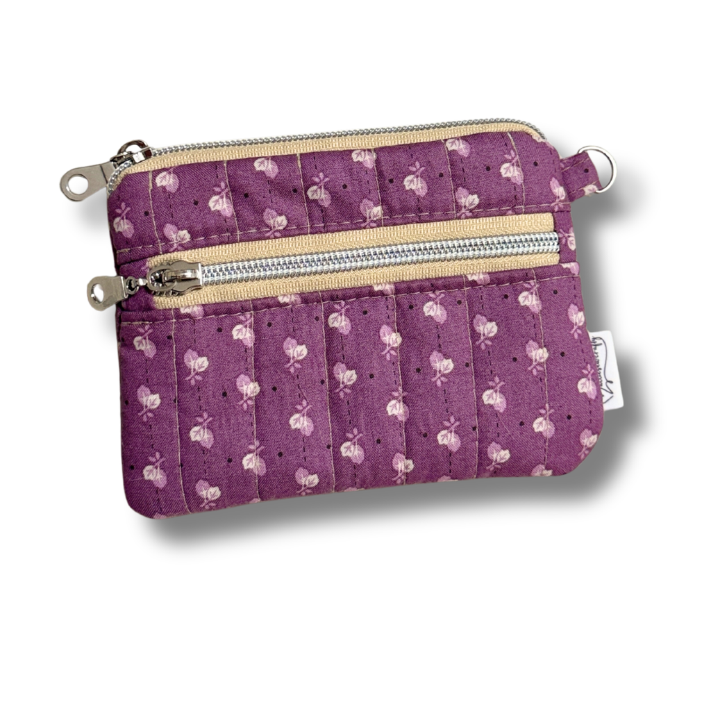 A purple quilted fabric wallet with a white floral pattern, featuring two zippered compartments, a card slot, and a keychain attachment in white background