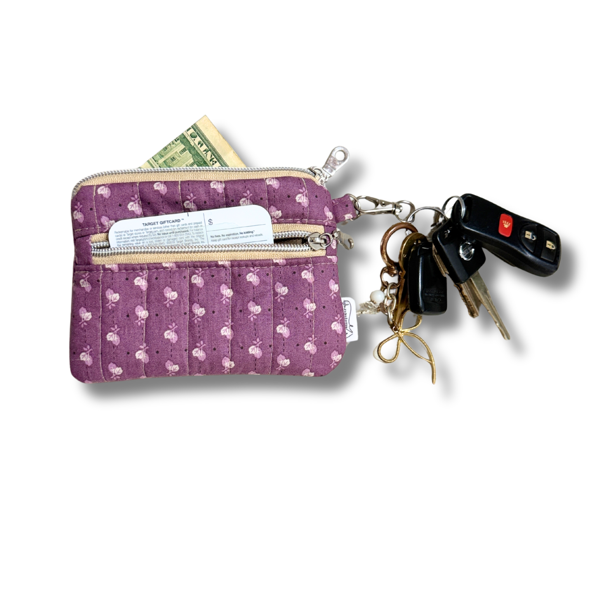 A purple quilted fabric wallet with a white floral pattern, featuring two zippered compartments, a card slot, and a keychain attachment with keys and a car key fob. A dollar bill is tucked into the top zipper.