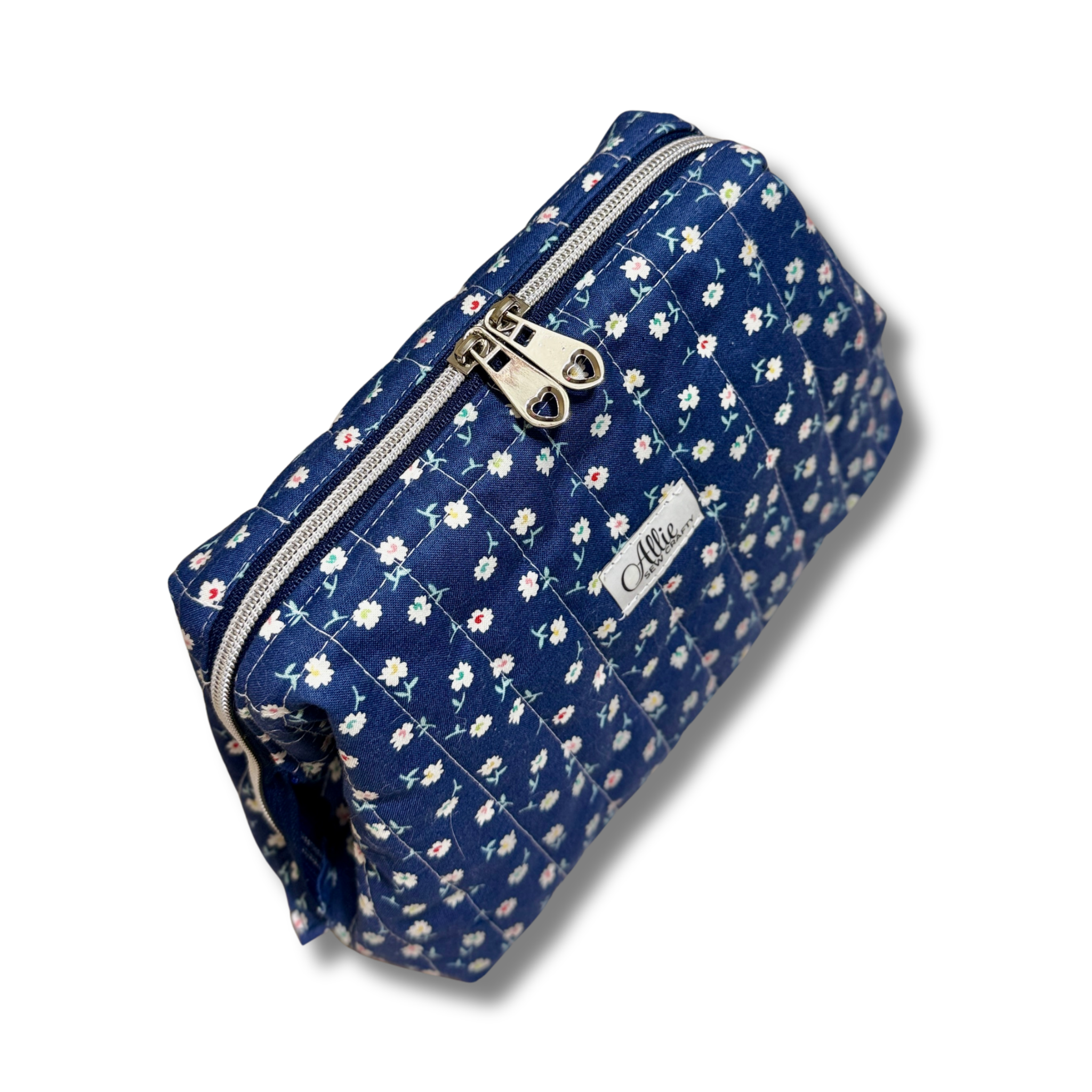 A navy blue quilted cosmetic pouch with a white and pink floral print, featuring a silver zipper and a white brand label that reads 'Allie Sew Crafty'