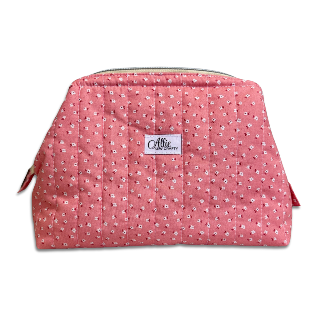 A pink quilted cosmetic pouch with a small white floral pattern, featuring a white brand label that reads 'Allie Sew Crafty'.