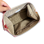 An open cosmetic pouch or makeup bag shown from the inside, with a hand holding the left side to display the interior. The pouch has a beige/tan interior lining, a silver zipper, and a pink patterned exterior visible at the edges.