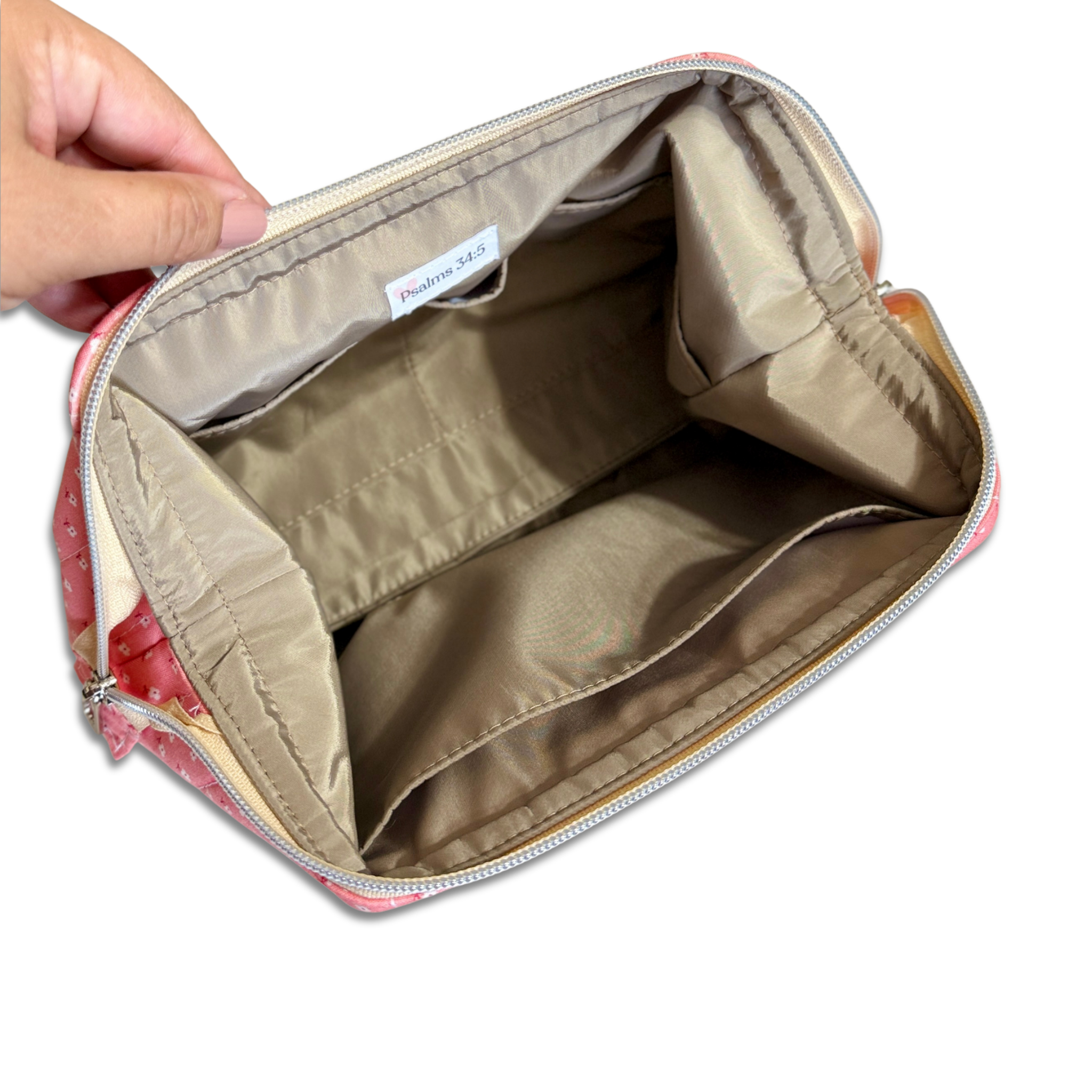 An open cosmetic pouch or makeup bag shown from the inside, with a hand holding the left side to display the interior. The pouch has a beige/tan interior lining, a silver zipper, and a pink patterned exterior visible at the edges.
