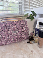 A floral makeup bag with a purple, pink, and white pattern sits on a marble countertop next to makeup brushes, a bottle of beauty spray, and a foundation bottle. A small white label on the front of the bag reads 'Affle.