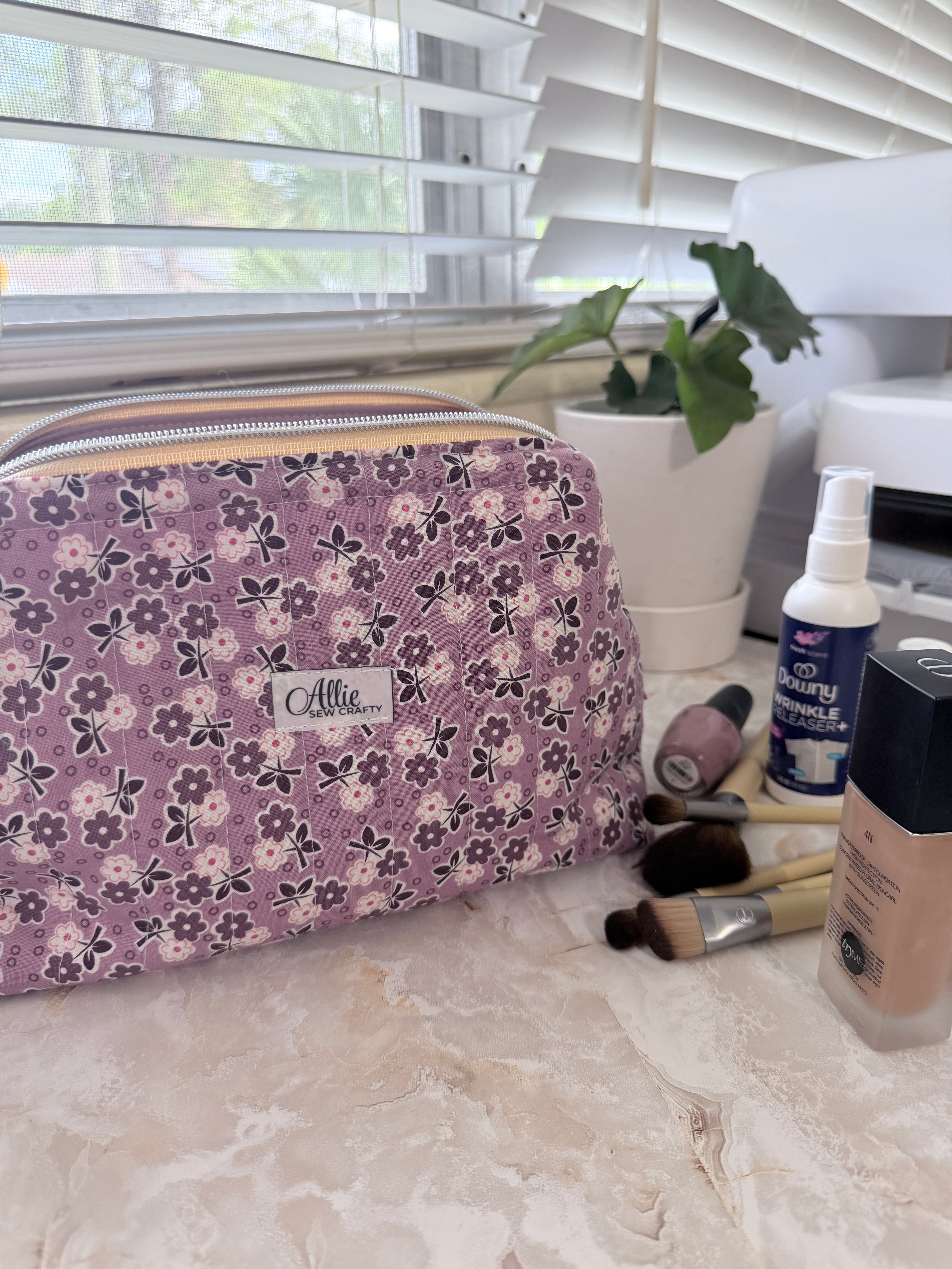 A floral makeup bag with a purple, pink, and white pattern sits on a marble countertop next to makeup brushes, a bottle of beauty spray, and a foundation bottle. A small white label on the front of the bag reads 'Affle.