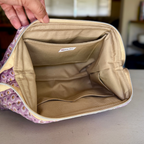 A close-up of a zippered cosmetic pouch being held open by a hand, showing a beige interior with multiple compartments and a small interior label. The exterior fabric has a lavender floral pattern.