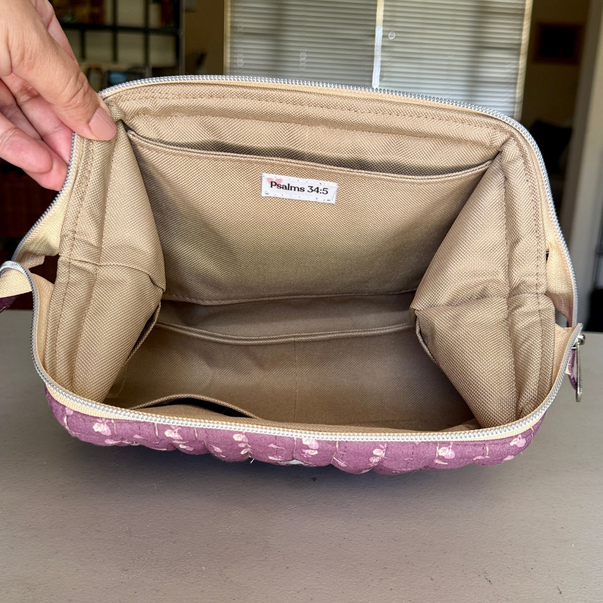 An open beige fabric organizer pouch with a pink patterned bottom edge, held by a hand. The interior features a white label reading 'Pistilna 34-5'.