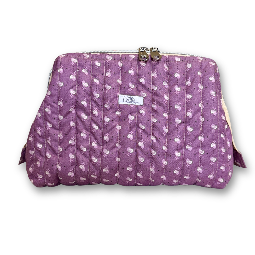 A quilted purple cosmetic pouch with a white and pink floral pattern, featuring a metal kiss-lock closure at the top and a small white brand label that reads 'Allie Sew Crafty".