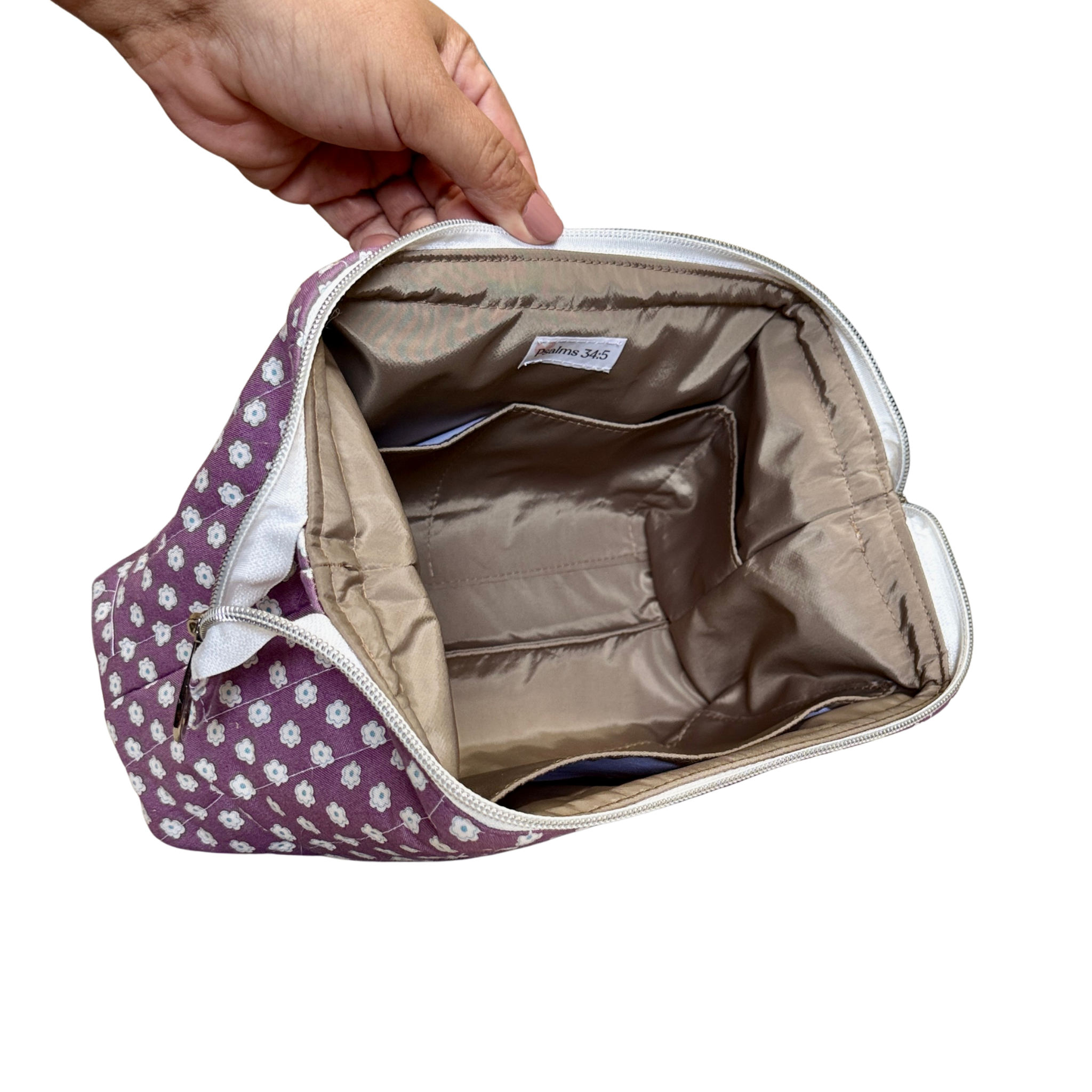 Hand opening a Purple White Daisies quilted Full View Pouch to show the tan water-resistant interior lining, interior pocket, and Psalm 34:5 tag