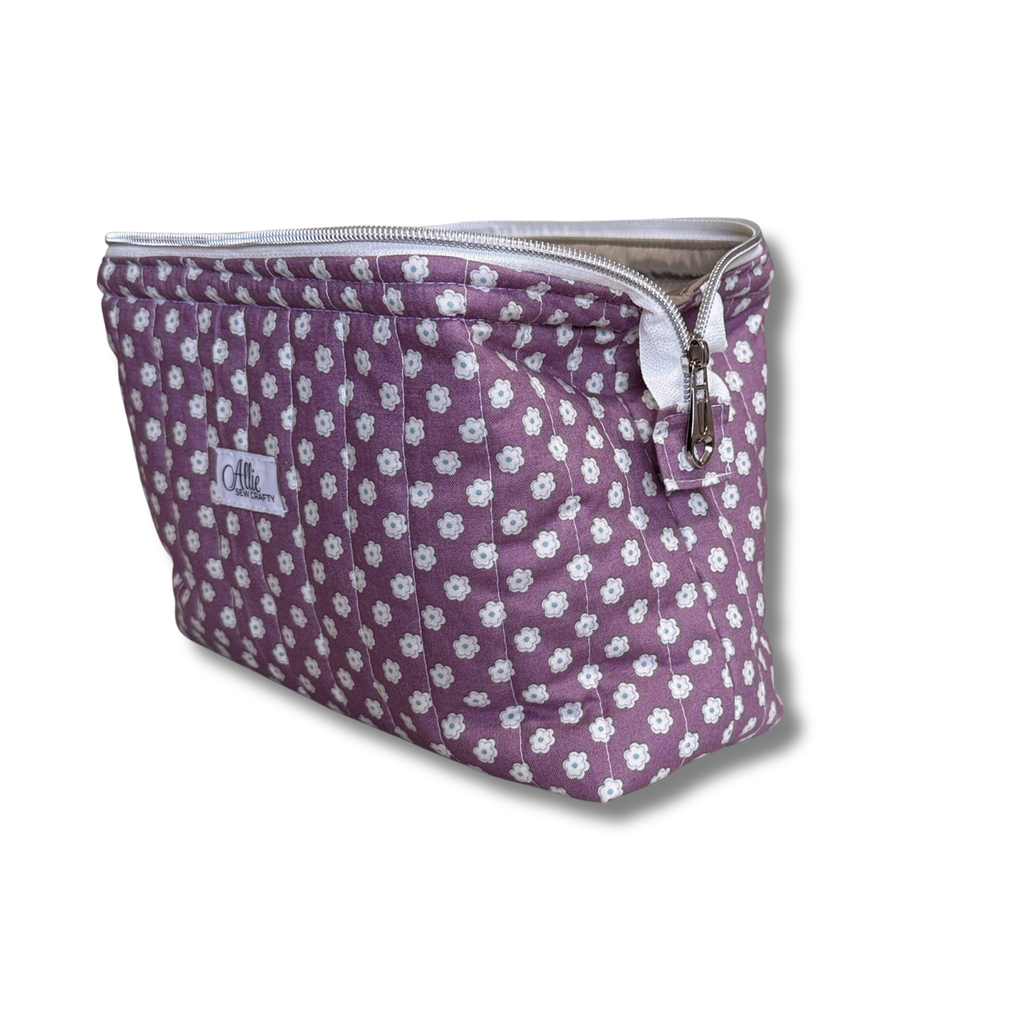 A quilted cosmetic pouch in a dusty purple fabric with a white floral pattern, featuring a silver zipper and a small embroidered brand label.