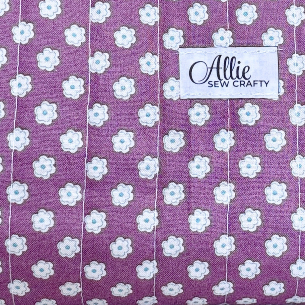 A close-up of a purple fabric with a repeating white daisy pattern, featuring a white sewn-on label that reads 'Allie Sew Crafty'.