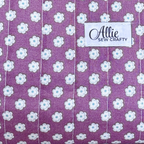 A close-up of a purple fabric with a repeating white daisy pattern, featuring a white sewn-on label that reads 'Allie Sew Crafty'.