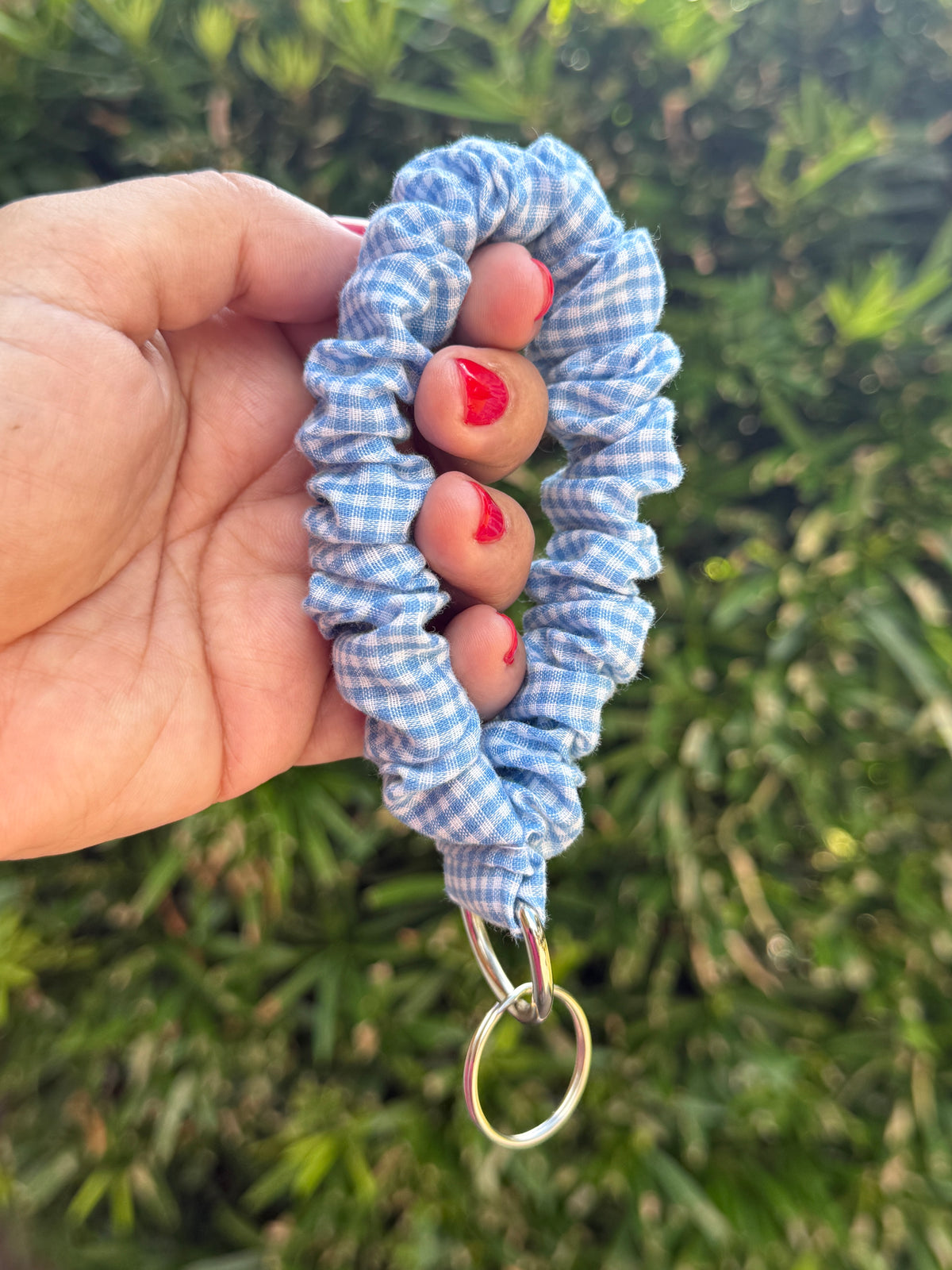 A hand holding a light blue and white gingham fabric scrunchie keychain with a silver metal split ring, set against a blurred green foliage background.