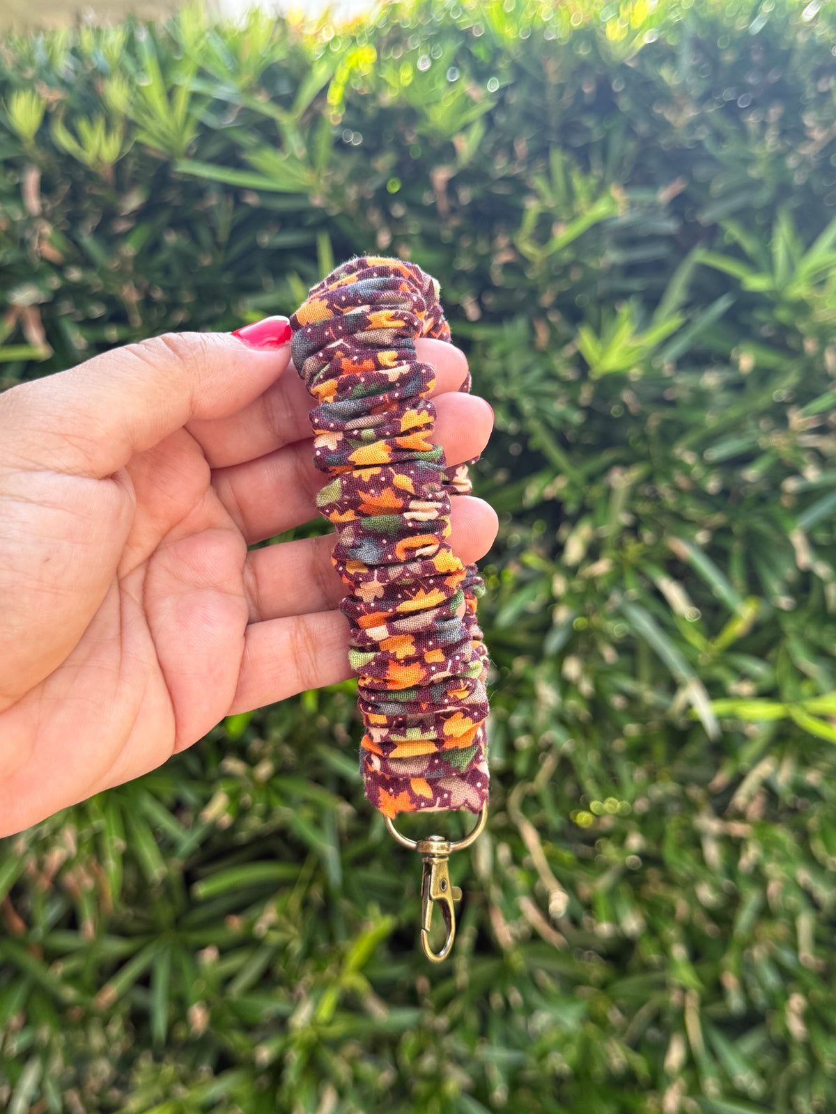 A hand holding a patterned fabric keychain or bag clip outdoors in front of green foliage. The fabric has a multicolor autumn pattern of orange, yellow, brown, and dark purple, with a gold-tone metal clasp at the bottom.