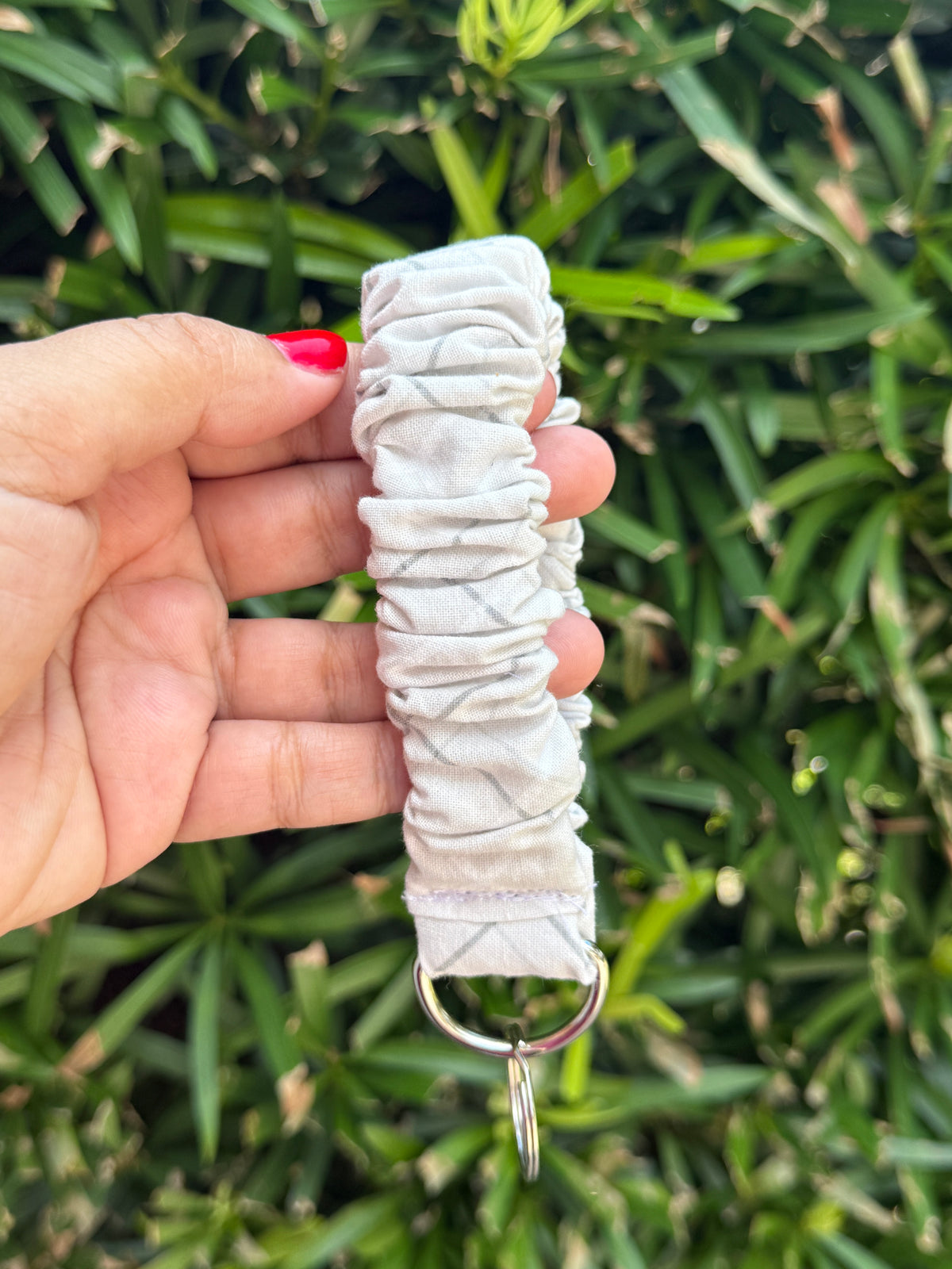 A hand holding a light gray fabric wrist strap or keychain loop with a metal ring, set against a background of green foliage.